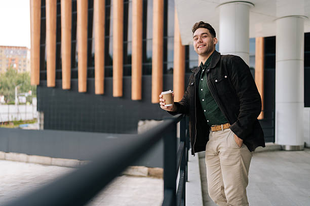 istock Vertical portrait of confident young man holding takeaway coffee cup leaning on railing while standing by modern building, smiling looking at camera. 2204435652