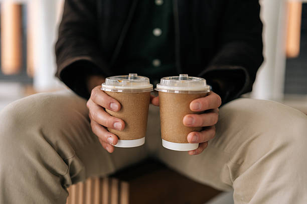 Close-up cropped shot of friendly man holding two disposable coffee cups, conveying warm gesture of sharing beverage in cozy