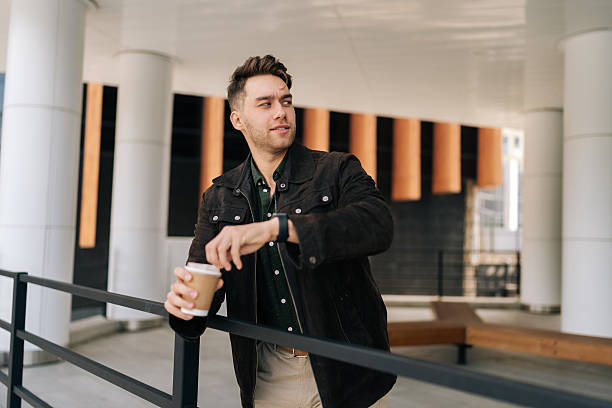 istock Bearded handsome young man enjoying coffee break, leaning against railing outside modern office building, smiling looking away at beautiful cityscape. 2204435666