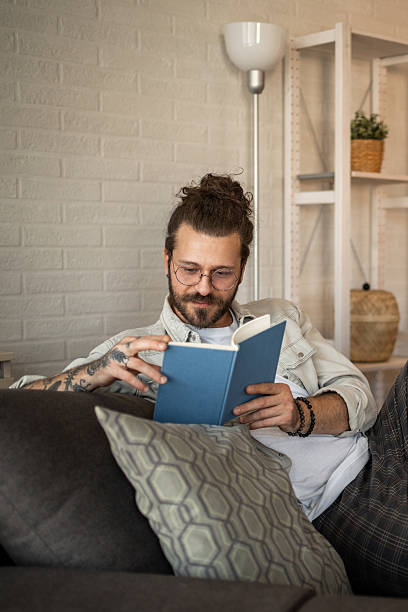 istock Young man reading a book on the sofa at home 2204437076