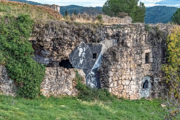 Ancient stone ruins of Montjuic Castle in Girona with white graffiti artwork decorating deteriorated walls and openings. Crumbling fortress structure overtaken by vegetation on grassy hillside. Dense forest and mountains visible in background under blue sky. Girona, Spain - November 28, 2021