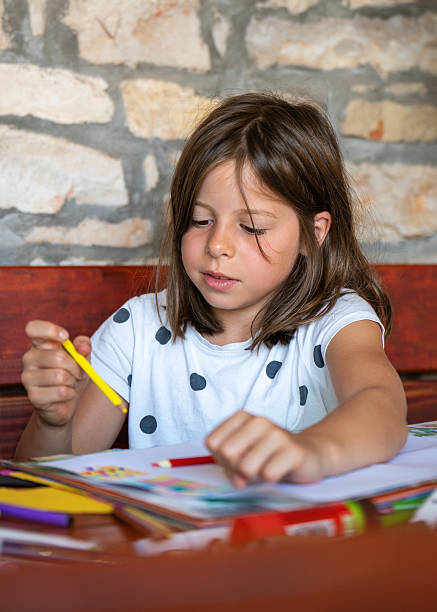 istock Enthusiastic girl draws picture while sitting at table in comfort her home. Bright colored pencils lie nearby. On childs face - concentration. Childhood, creativity, development imagination. 2204448117