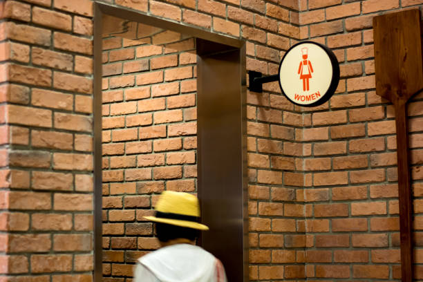 A person wearing a hat approaches a restroom sign in a brick-walled interior.
