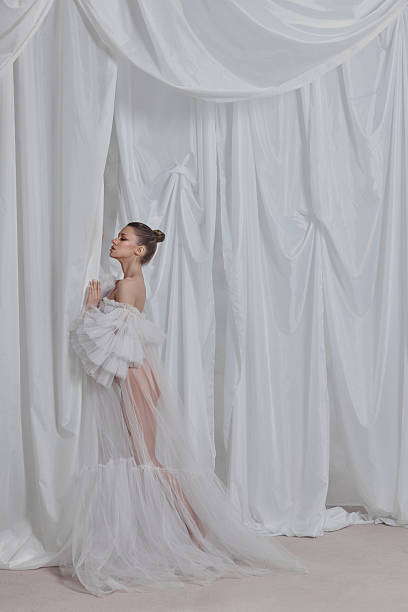 istock Model stands gracefully in a stunning white gown, leaning against a soft draped fabric backdrop. setting highlights the delicate fabric and the models elegant pose during a fashion shoot 2204470660