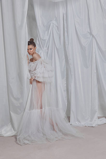 istock woman showcases a delicate white gown with layered ruffles, striking a pose beside elegant drapery. Soft lighting accentuates the couture design in a serene studio atmosphere 2204470664