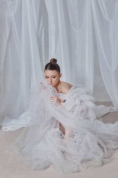 istock model sits elegantly on the floor, surrounded by sheer white drapery. She wears a delicate, flowing outfit that hints at softness and grace, creating a serene and artistic atmosphere 2204470665