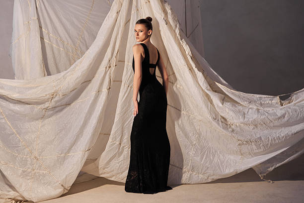 model showcases an elegant black gown in a studio adorned with flowing fabric. lighting highlights the gowns intricate details and the models confident pose captures attention
