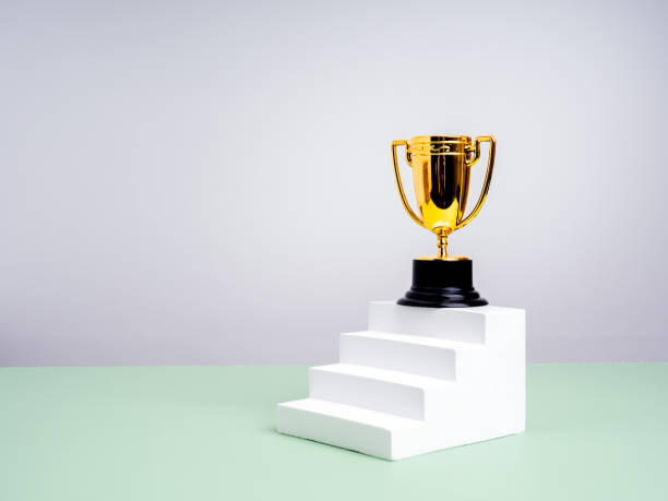 istock Gold trophy on top of white stair steps as bar graph, on green table, white background with copy space. Business goal and success, achievement, competition, leadership mission, career growth concepts. 2204472914