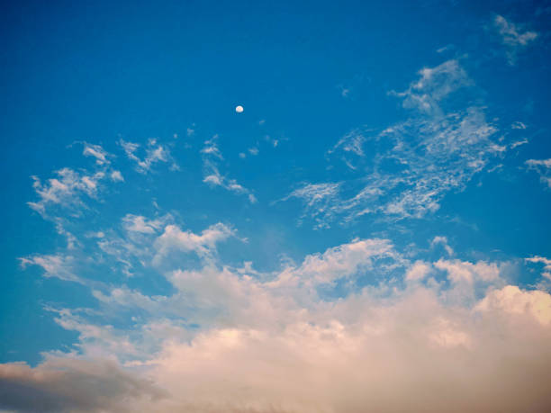 Bright blue sky with wispy white clouds and a small moon.  Lower clouds have warm reddish tones suggesting sunrise or sunset.