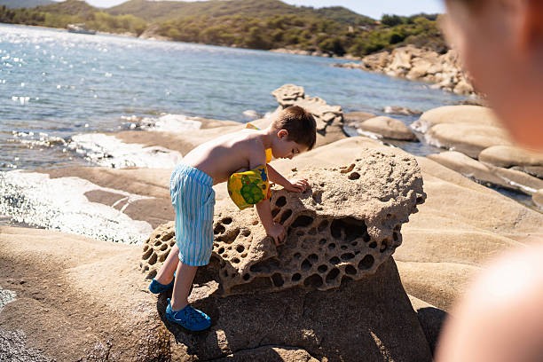Little boy playing on the rocky beach while his mother watching on him.