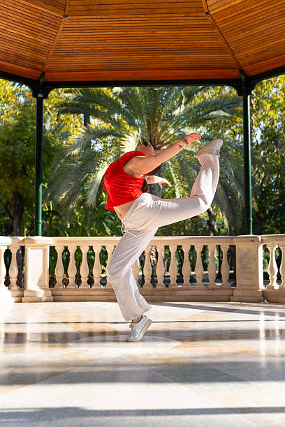 Young woman performing fitkid acrobatics in a gazebo Flexible athlete practicing fitkid, jumping and dancing gracefully in a gazebo, showcasing strength and elegance in a vibrant park setting power balance pavillion stock pictures, royalty-free photos & images