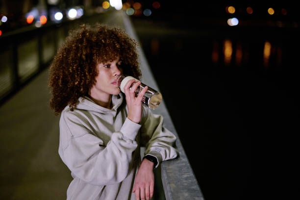 Young woman drinking water from a reusable bottle after night exercising, leaning on a bridge railing, with city lights blurred in the background
