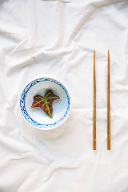 A beautifully arranged minimalist composition featuring a traditional blue and white ceramic bowl with a single autumn leaf inside, placed on soft white fabric. Alongside the bowl, a pair of wooden chopsticks creates a balanced and harmonious layout. The simplicity of this image highlights themes of mindfulness, nature, and artistic presentation, making it ideal for conceptual photography, food styling, cultural design projects, and visual storytelling focused on organic aesthetics.