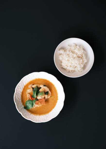 Traditional Thai curry with chicken and vegetables served in a white bowl, accompanied by steamed white rice in a separate dish, placed on a dark background, Authentic Asian cuisine concept