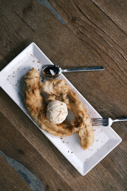 istock Top view of Thai fried banana dessert with vanilla ice cream, served on a white rectangular plate with spoon and fork, rustic wooden table setting, classic Asian sweet treat 2204488911