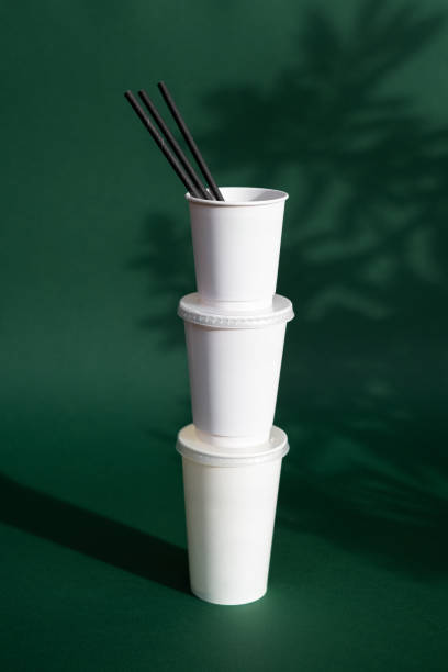 Stack of white takeaway cups with black straws against a green background, concept of sustainable packaging, eco-friendly food containers, and modern beverage culture