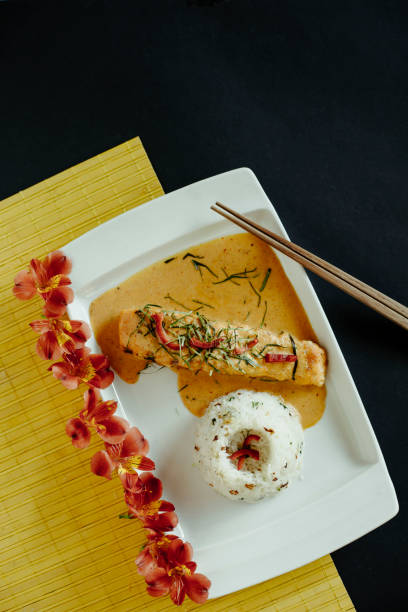 A beautifully plated Thai-inspired dish featuring a tender salmon fillet covered in a rich and creamy red curry sauce, accompanied by a neatly shaped portion of jasmine rice. The plate is decorated with delicate edible flowers, adding an artistic and luxurious touch to the presentation. The dish is served on a contrasting black and yellow placemat with chopsticks, enhancing the vibrant aesthetic. This image perfectly showcases fusion cuisine, fine dining, and modern Thai gastronomy, making it ideal for restaurant branding, food photography, and gourmet culinary presentations.