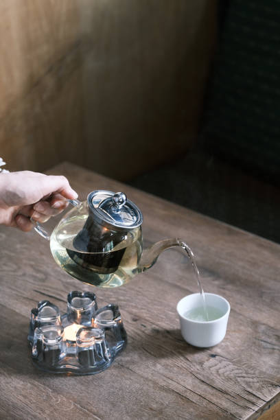 istock Glass teapot pouring herbal tea into a white cup on a rustic wooden table, Warm and cozy tea ceremony with natural lighting, Relaxing moment of mindfulness and traditional tea culture 2204489968