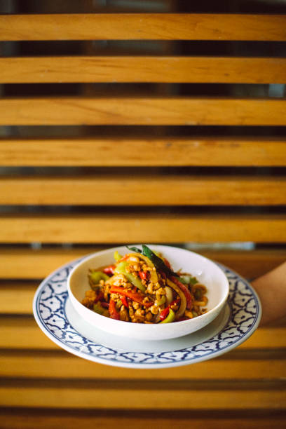 A visually striking image featuring a hand presenting a beautifully plated serving of Pad Krapow Gai, a classic Thai stir-fried basil chicken dish. The vibrant colors of the dish, including red chilies, fresh basil, and golden-brown chicken, contrast with the traditional blue and white ceramic plate. The modern wooden slat background adds depth and texture, blending contemporary and cultural aesthetics. This image represents the rich flavors of Thai street food, gourmet presentation, and the warmth of traditional Asian hospitality.