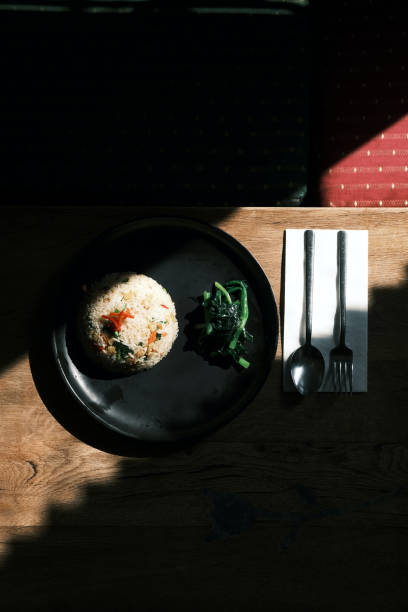 A beautifully composed top-down shot of Thai fried rice elegantly plated on a black dish, accompanied by fresh greens. The meal is set on a rustic wooden table with natural light casting strong shadows, creating a striking contrast that enhances the textures and colors of the dish. A neatly placed spoon and fork with a white napkin add a refined touch to the presentation. This image perfectly captures the essence of authentic Thai cuisine, making it ideal for restaurant menus, food blogs, and culinary storytelling.