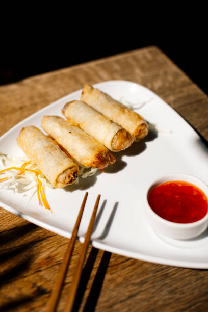 istock Top view of crispy golden spring rolls served on a white triangular plate with a side of red sweet chili dipping sauce and chopsticks, placed on a rustic wooden table under dappled sunlight, Asian appetizer concept 2204490645