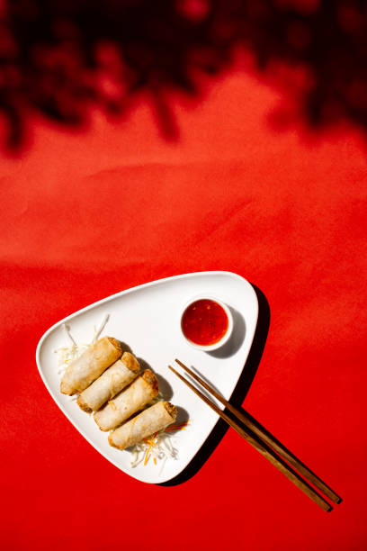 istock Top view of crispy golden spring rolls arranged on a white triangular plate with a side of red sweet chili dipping sauce and wooden chopsticks, set against a vibrant red background with artistic shadows, Asian appetizer concept 2204490679