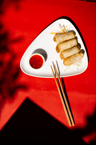 istock Top view of crispy golden spring rolls arranged on a white triangular plate with a side of red sweet chili dipping sauce and wooden chopsticks, set against a vibrant red background with artistic shadows, Asian appetizer concept 2204490682