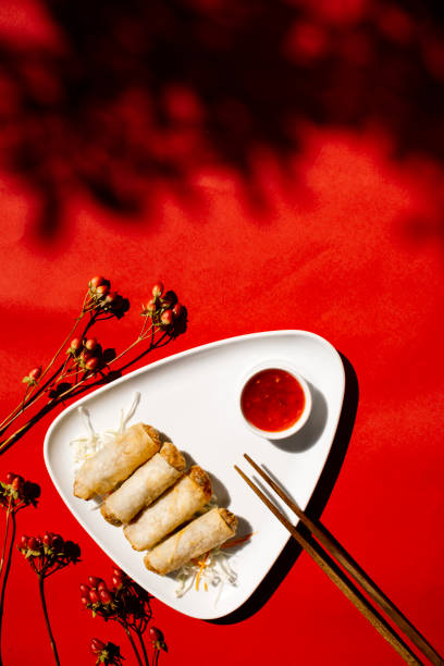 Top view of golden crispy spring rolls arranged on a white triangular plate with a side of red sweet chili sauce and wooden chopsticks, set against a vibrant red fabric background with delicate dried flowers and artistic shadows, Asian appetizer concept