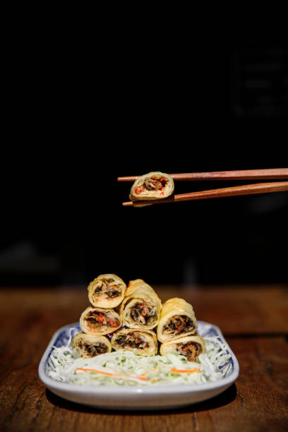 A perfectly balanced stack of golden, crispy spring rolls served on a blue and white ceramic plate, placed on a rustic wooden table. One piece is delicately lifted by wooden chopsticks, showcasing the delicious filling inside. The rolls are arranged on a bed of finely shredded cabbage, enhancing their visual appeal. The dark moody background contrasts beautifully with the warm tones of the fried rolls, making it ideal for food photography, Asian restaurant branding, and gourmet culinary presentations.
