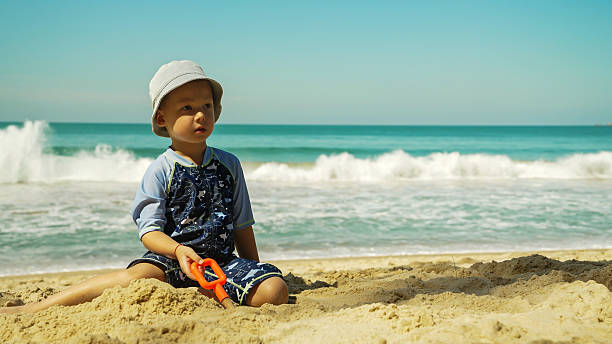 A young child sits on warm sand at the beach, focusing intently on a toy shovel. The clear blue ocean waves crash gently in the background under a bright sky.
