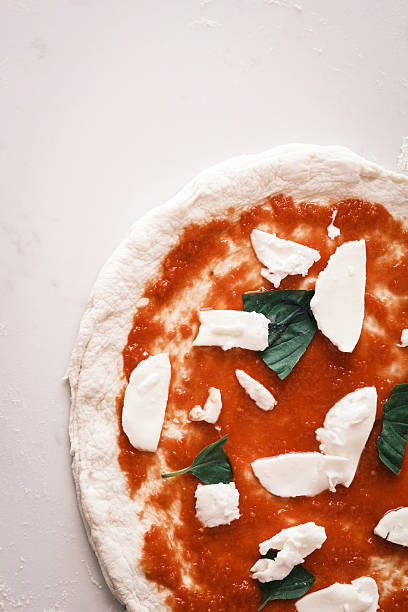 Top view of uncooked Neapolitan-style pizza with tomato sauce, fresh mozzarella chunks and basil leaves on raw dough, Minimal food styling on neutral background, Traditional Italian cuisine preparation concept