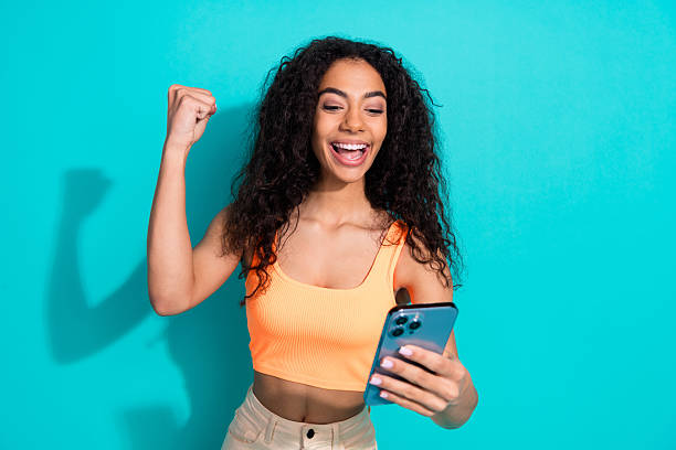 istock Excited young woman celebrating while using her smartphone against a turquoise background, wearing stylish casual summer attire 2204502107