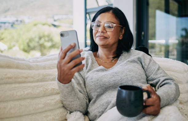 Mature African Woman Sitting on Sofa, Holding Coffee Mug, and Using Mobile Phone stock photo