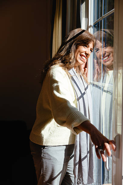 istock Smiling middle-aged woman looking out the window with sunlight reflection 2204517701
