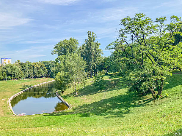 istock Image shows a serene park setting with a calm water body reflecting the clear sky, gentle green slopes, and mature deciduous trees People are walking or standing in the background 2204521974