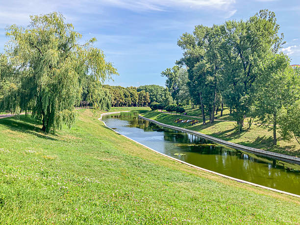 A serene park scene showcases a curved path, small riverpond, lush green grass, well-maintained ground, and various tree species against a clear blue sky