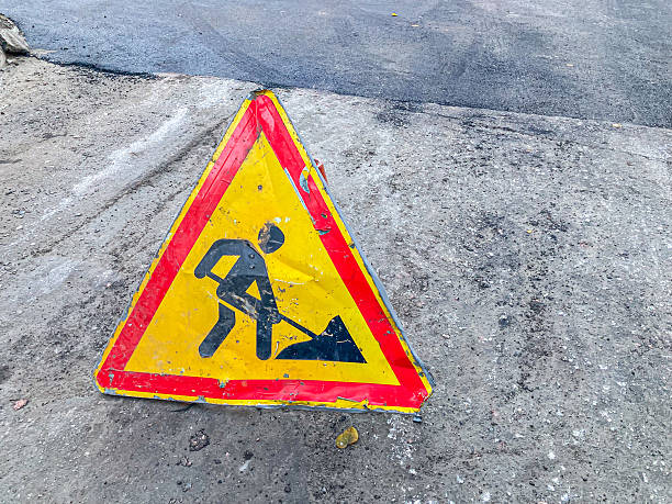 istock A blurry image shows a warning sign of construction or roadwork, symbolized by a shovel-wielding figure The sign leans against a curb or boundary, possibly near a roadway with pavement and grass 2204525461