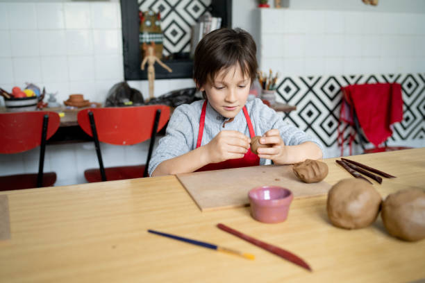 A young boy is sitting at a work table as he is molding clay for a creative project during a pottery workshop at school