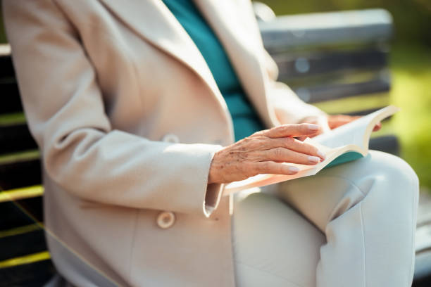 A closeup view of an older woman wearing a jacket while reading a book outdoors on a sunny day, providing a sense of calm and leisure.