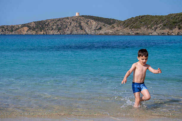 A young boy is running in the sea A young boy is running in the sea. The water is blue and the sky is clear. The boy is wearing a blue swimsuit and is barefoot. The sea is a sardinian beach called Tuerredda costa esmeralda stock pictures, royalty-free photos & images