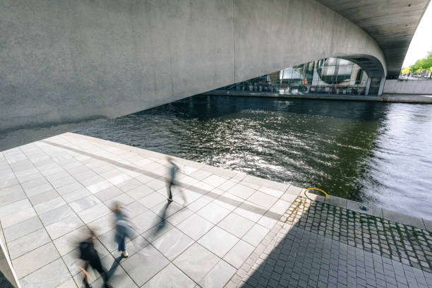 A concrete bridge spans a tranquil urban river. Pedestrians walk along a sunny pathway by the water.\nGovernment District in Berlin