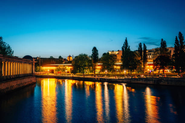 The photo captures a vibrant twilight scene in Berlin, Germany, with city lights reflecting on a serene river, surrounded by historic architecture and trees during an enchanting evening.