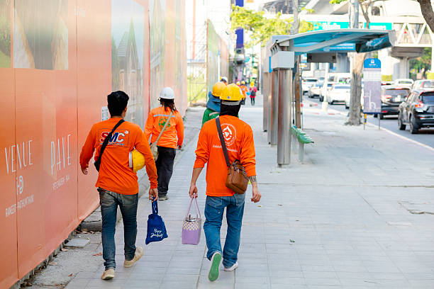 Rear view of walking group of construction workers leaving construction site in Bangkok. They are walking  on sidewalk of Phahonyothin Road in Bangkok Chatuchak. People are passing a huge banner at fence of construction site. Some are wearing safety helmet. They carry bags for food etc. In background is a bus stop. At right side is traffic jam on Phahonyothin Road
