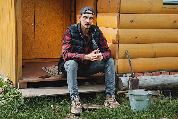 A young hipster sitting on the terrace bench with his legs leaning against a wooden pole and drinking coffee melancholic autumn mood