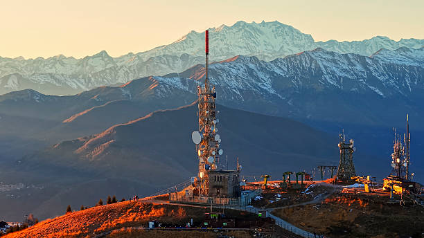 Panoramic view of telecommunications infrastructure silhouetted against snow-covered Alpine peaks at Monte Mottarone. Golden sunlight illuminates mountain ridges during sunset in northern Italy.