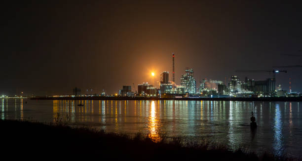 Oil refinery and illuminated industrial plant situated along a large river. A tall chimney releasing a fuel flare
