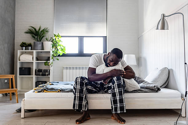 A young man in cozy pajamas sits on his bed, hugging a pillow with a thoughtful expression. The modern bedroom setting and soft lighting create a peaceful, introspective atmosphere.