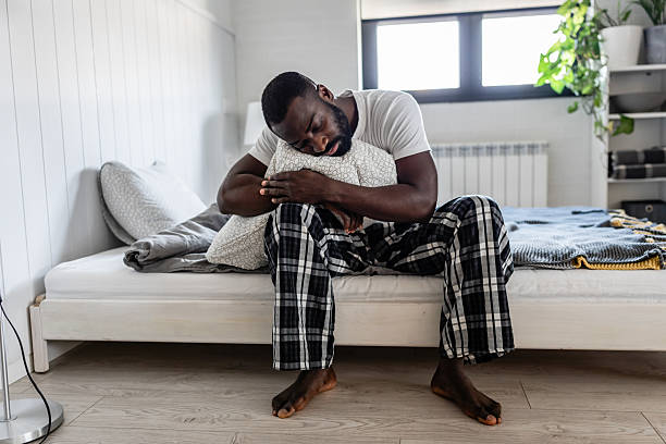 A young man in cozy pajamas sits on his bed, hugging a pillow with a thoughtful expression. The modern bedroom setting and soft lighting create a peaceful, introspective atmosphere.