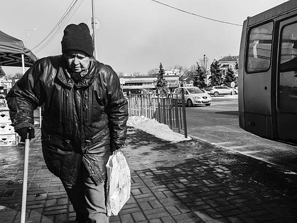 Kyiv, Ukraine - 2d March, 2025: An elderly man walks carefully along the sidewalk, clutching a bag of groceries, as the chilly breeze sweeps through the bustling city.
