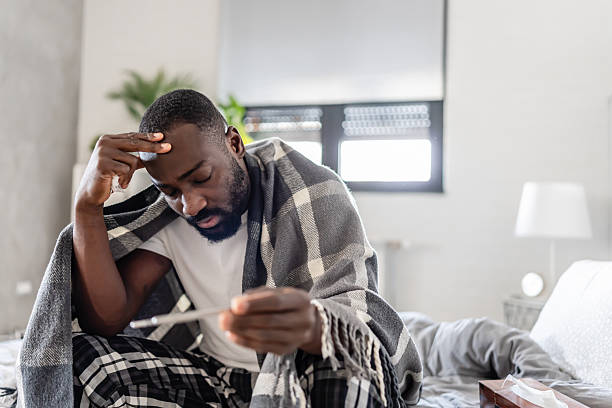 istock A sick man wrapped in a blanket sits at home, holding a thermometer and touching his forehead. He looks unwell and concerned, possibly dealing with fever or illness. 2204567170